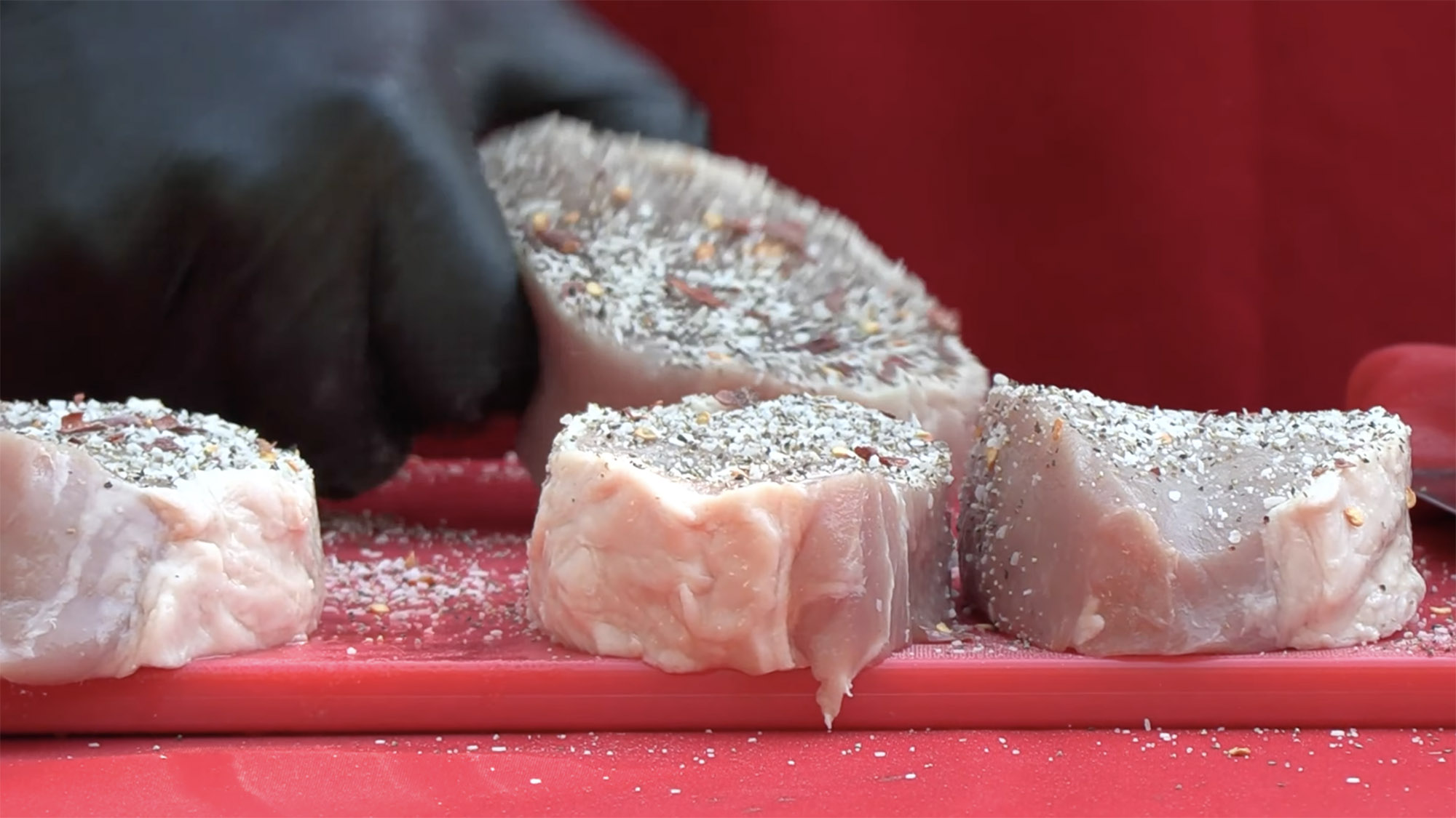 Close shot of raw pork chops on a red tabletop with a dry rub mix sprinkled over them for cooking.